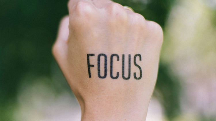 A close-up of a hand with the word 'FOCUS' tattooed in bold black letters on the wrist, symbolizing mindfulness and concentration against a blurred green background