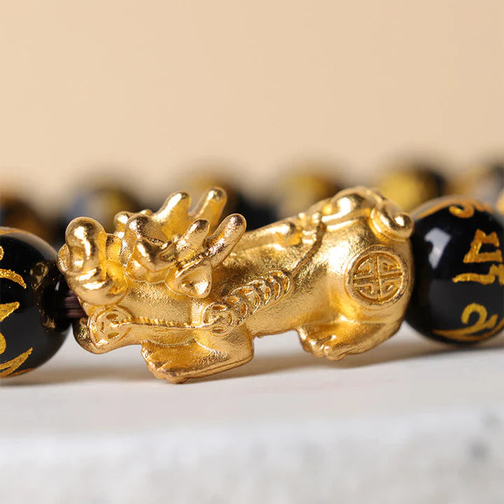 Detailed macro shot of an engraved golden Pixiu charm on a natural black obsidian beaded bracelet.